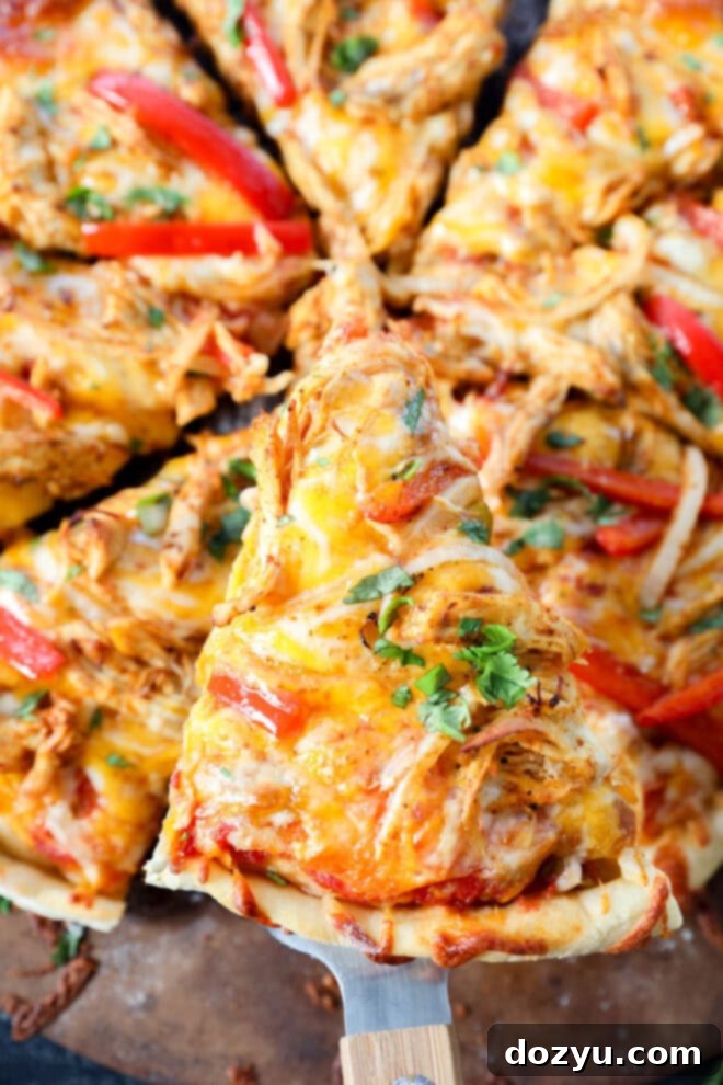 Sizzling Fajita Pizza 7 A close-up of a pizza topped with shredded chicken, melted cheese, red bell peppers, onions, and fresh herbs. One slice is being lifted from the whole chicken fajita pizza with a spatula.