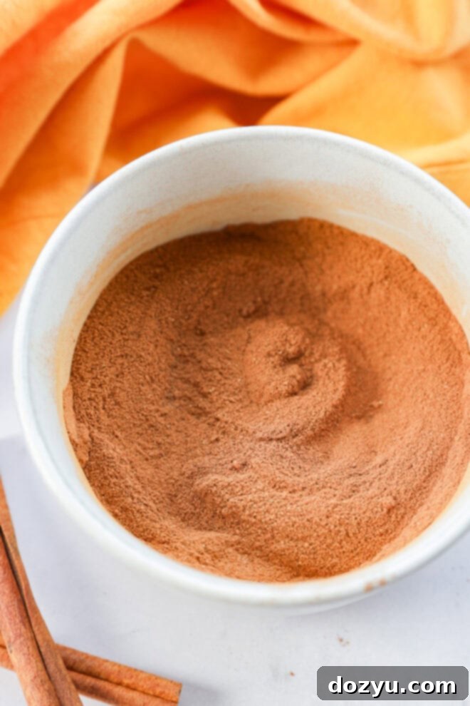 A white bowl filled with a mound of rich, golden-brown homemade pumpkin pie spice, with natural cinnamon sticks, a whole nutmeg, and an orange linen cloth arranged artfully on a light wooden surface, evoking the warmth of fall.
