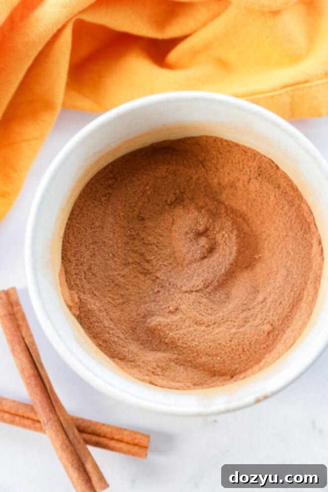 A beautifully textured white ceramic bowl brimming with a generous mound of rich, finely ground homemade pumpkin pie spice. Two natural cinnamon sticks lie beside the bowl on a pristine white surface, with a soft orange cloth partially visible in the background, highlighting the cozy essence of fall.