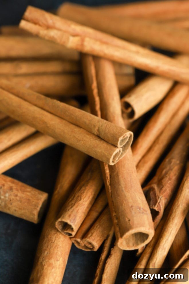 A rustic pile of smooth, reddish-brown cinnamon sticks, varying in length and thickness, with a few lighter pieces, arranged on a light surface, ready to impart warmth and fragrance.