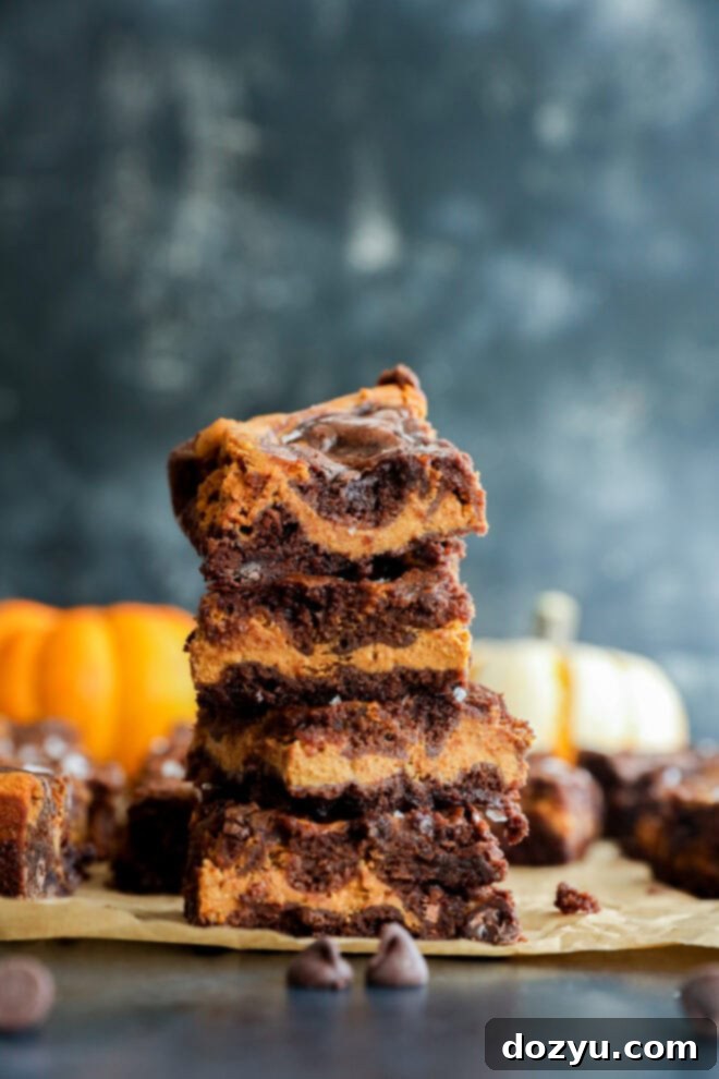 A delectable stack of four swirled chocolate and pumpkin bars rests on parchment paper, adorned with scattered chocolate chips, against a blurred backdrop of autumnal pumpkins, signaling a cozy fall treat.