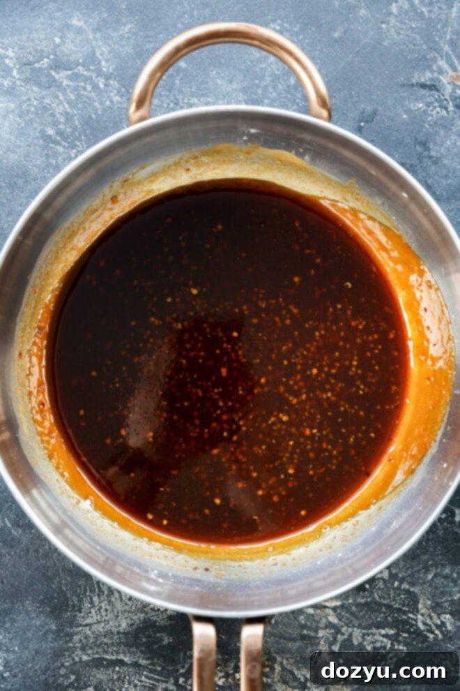 Sweet and Sticky Honey Garlic Chicken Wings 5 A saucepan filled with a dark brown sauce, likely caramel or soy-based, sits on a textured gray surface. The sauce has a glossy appearance with small specks throughout and some residue on the pans sides.