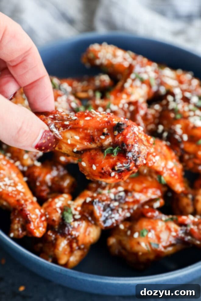 Sweet and Sticky Honey Garlic Chicken Wings 8 A close-up of a hand picking up a glazed chicken wing from a blue bowl filled with sticky, saucy wings garnished with white sesame seeds and chopped herbs.