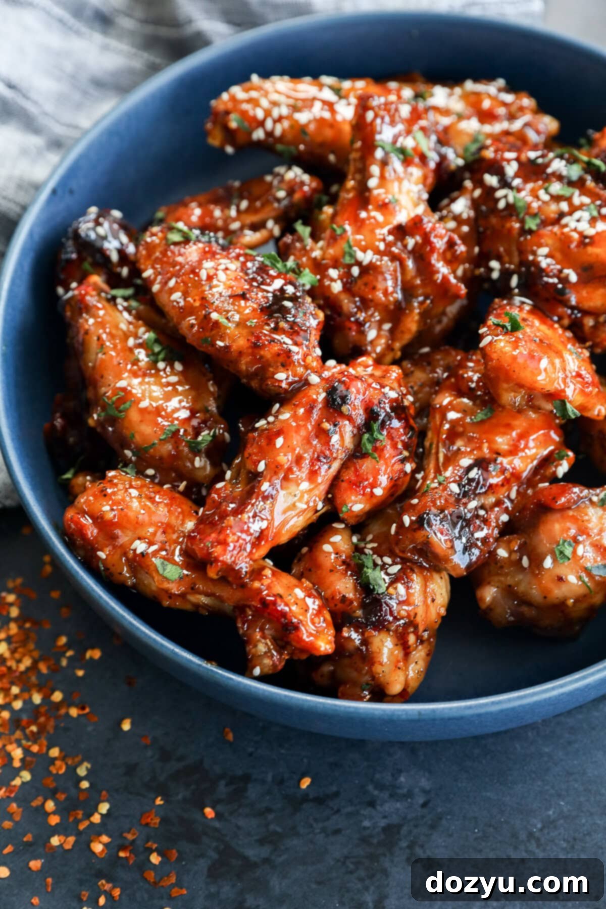 Sweet and Sticky Honey Garlic Chicken Wings 10 A blue bowl filled with glazed chicken wings sprinkled with sesame seeds and chopped herbs, with some red pepper flakes scattered on a dark surface nearby.