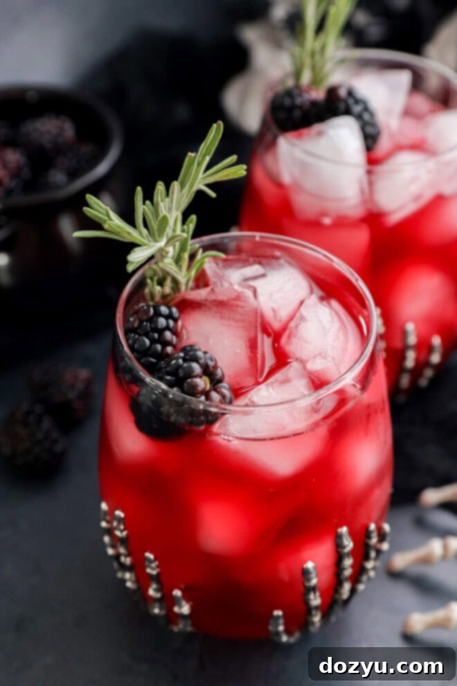 A glass of witches brew over ice, garnished with blackberries and a sprig of rosemary. Another similar drink is in the background, both served in beaded glasses on a dark surface.