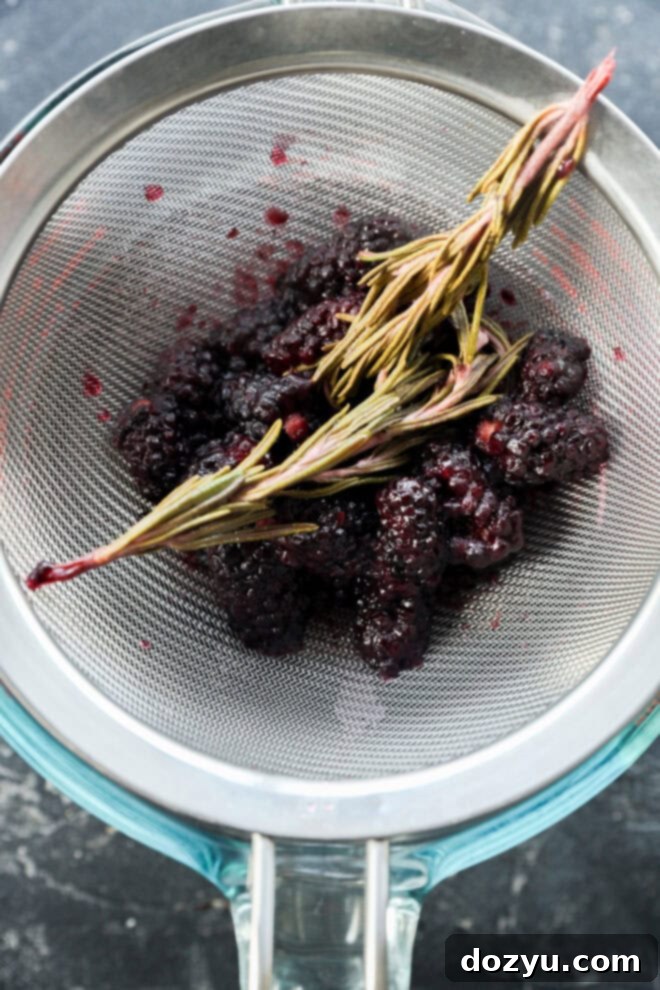 A metal strainer holds mashed blackberries and sprigs of rosemary over a glass measuring cup on a dark surface. Some juice stains are visible on the strainer.