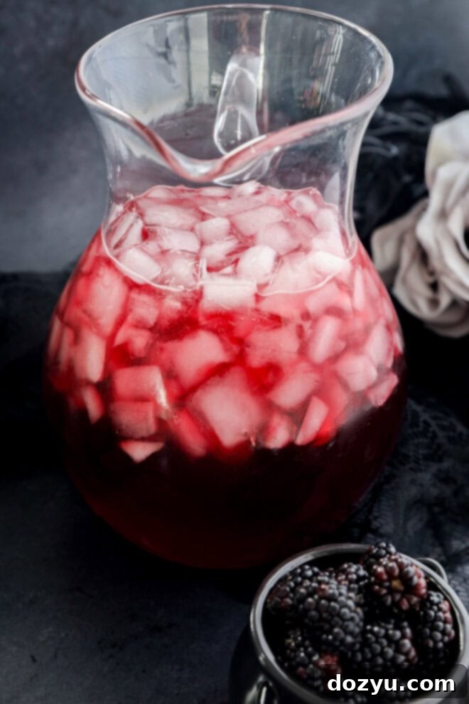A glass pitcher filled with a red beverage and ice cubes sits on a dark surface. Nearby, a small bowl holds fresh blackberries. A white cloth is partially visible in the background.