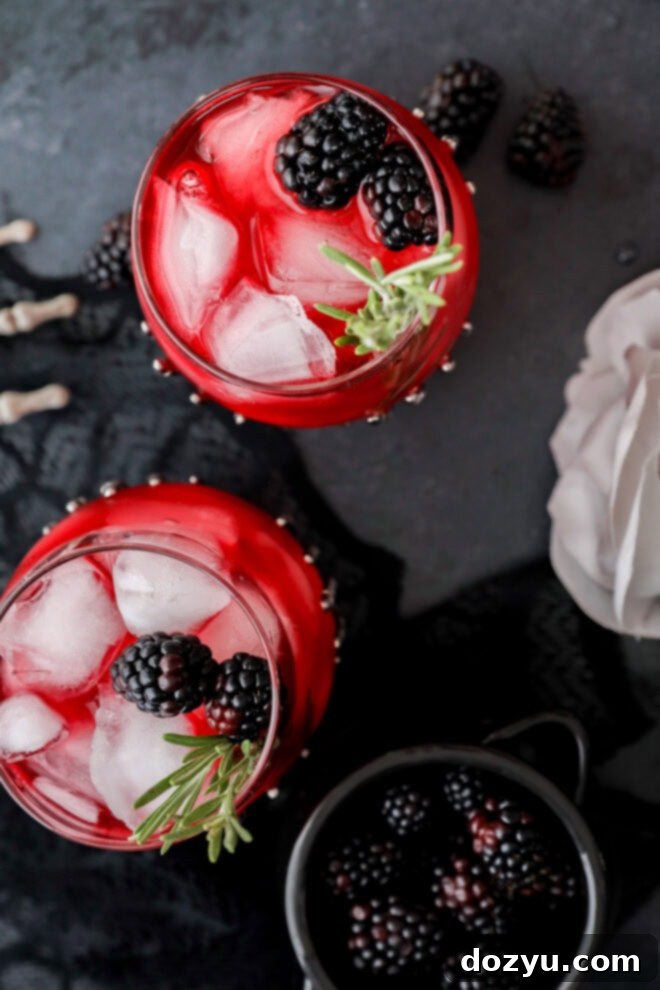 A top-down view of a witches brew—a red drink with ice, fresh blackberries, and a sprig of rosemary in a glass, with some blackberries scattered nearby on a dark surface.