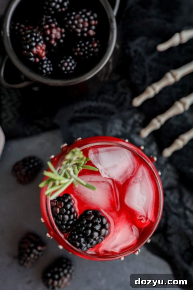 A red drink with ice cubes, blackberries, and a sprig of rosemary in a glass, with loose blackberries scattered nearby, a metal bowl of blackberries, and a decorative skeleton hand on a dark surface.