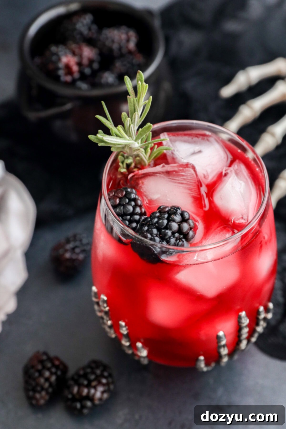A Witches Brew glass with ice cubes is garnished with blackberries and a rosemary sprig in a decorated glass. A small bowl of blackberries and a skeleton hand prop are in the background, creating a spooky atmosphere.