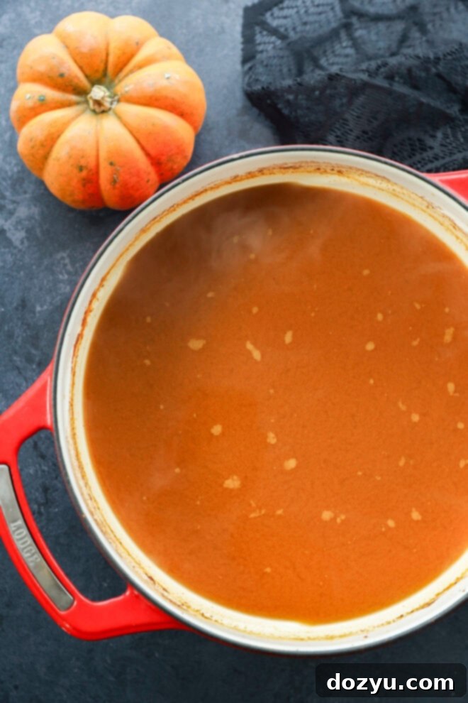 A red Dutch oven filled with smooth, orange fall beverage sits on a dark surface next to a small pumpkin and a piece of black fabric.