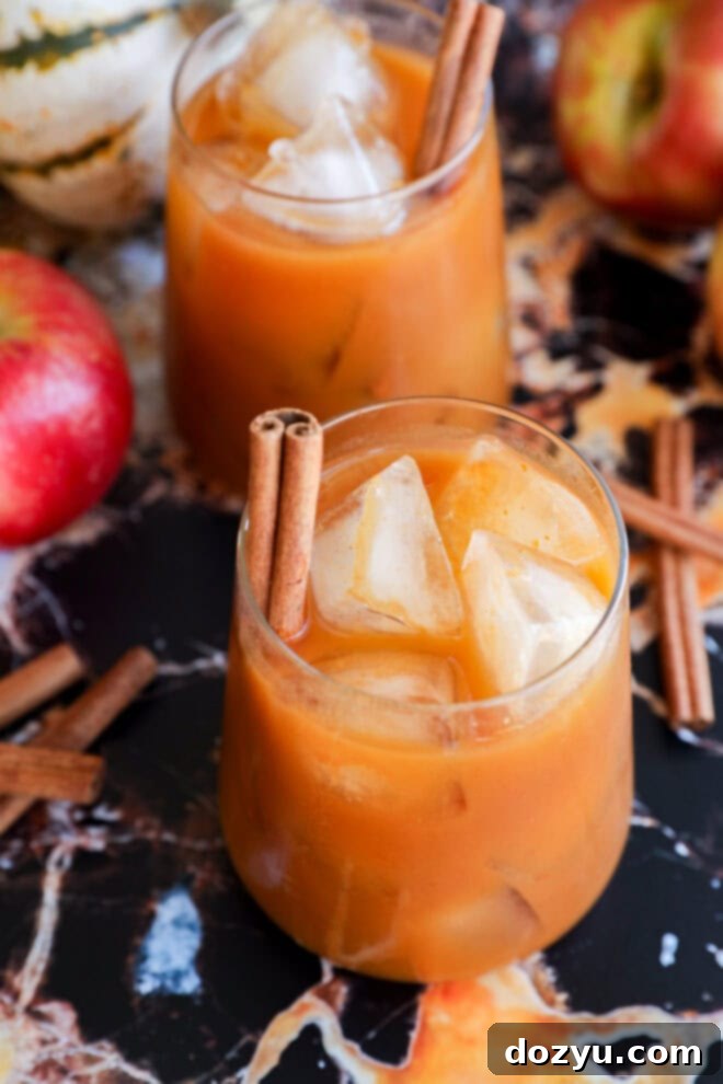 Two glasses of iced apple cider garnished with cinnamon sticks are on a marble surface, surrounded by fresh apples, loose cinnamon sticks for a cozy autumn twist.