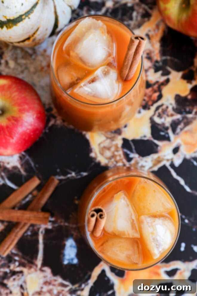 Two glasses of iced pumpkin juice, garnished with cinnamon sticks, sit on a marbled surface surrounded by apples, a small white pumpkin, and loose cinnamon sticks.