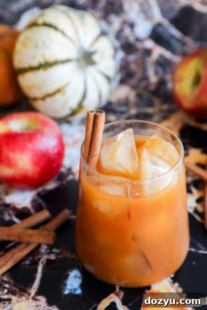 A glass of orange iced pumpkin juice garnished with two cinnamon sticks, surrounded by apples, extra cinnamon sticks, and small decorative pumpkins on a marble surface.