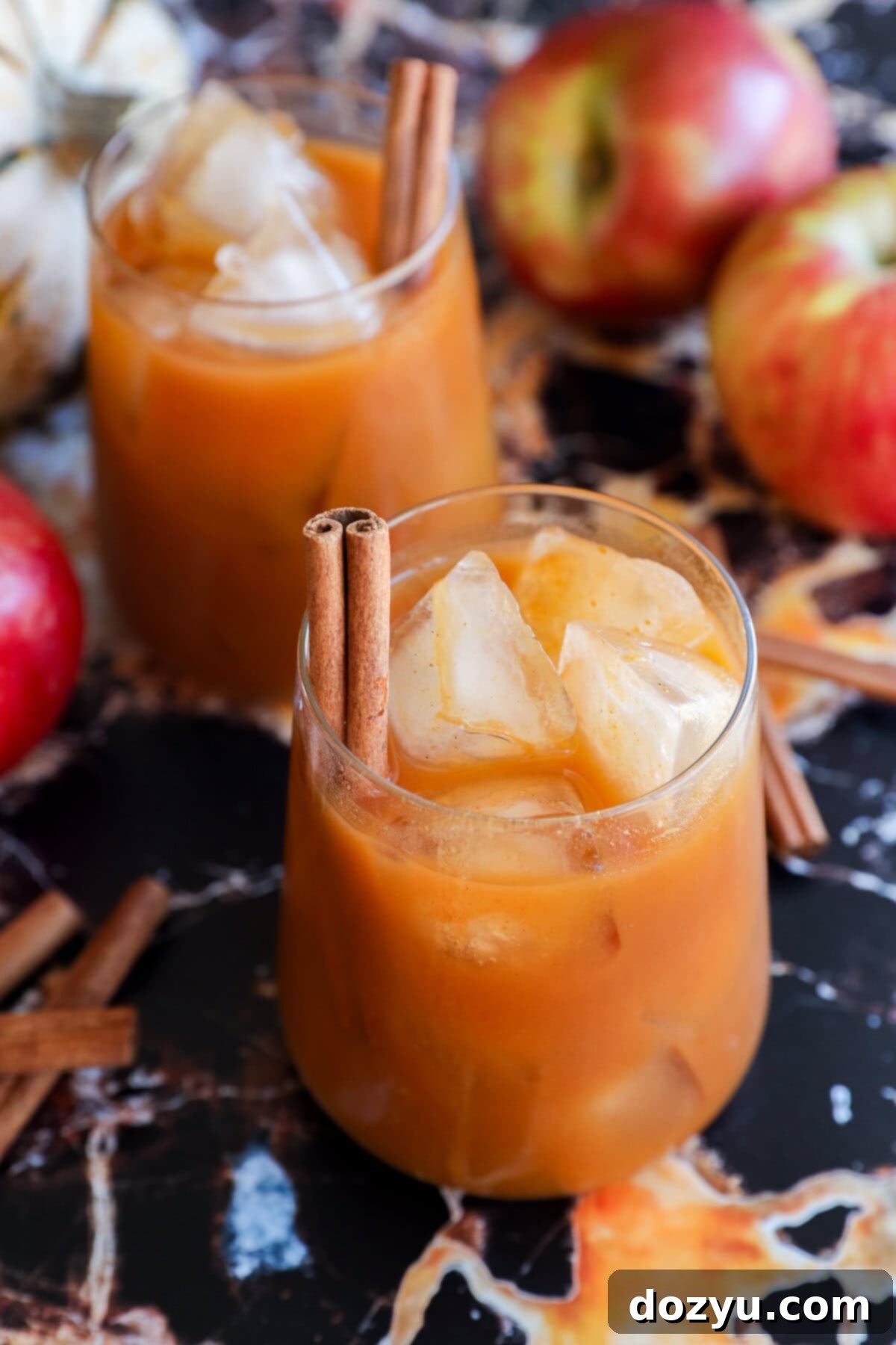 Two glasses filled with pumpkin juice, garnished with cinnamon sticks, sit on a dark marble surface surrounded by red apples and extra cinnamon sticks.