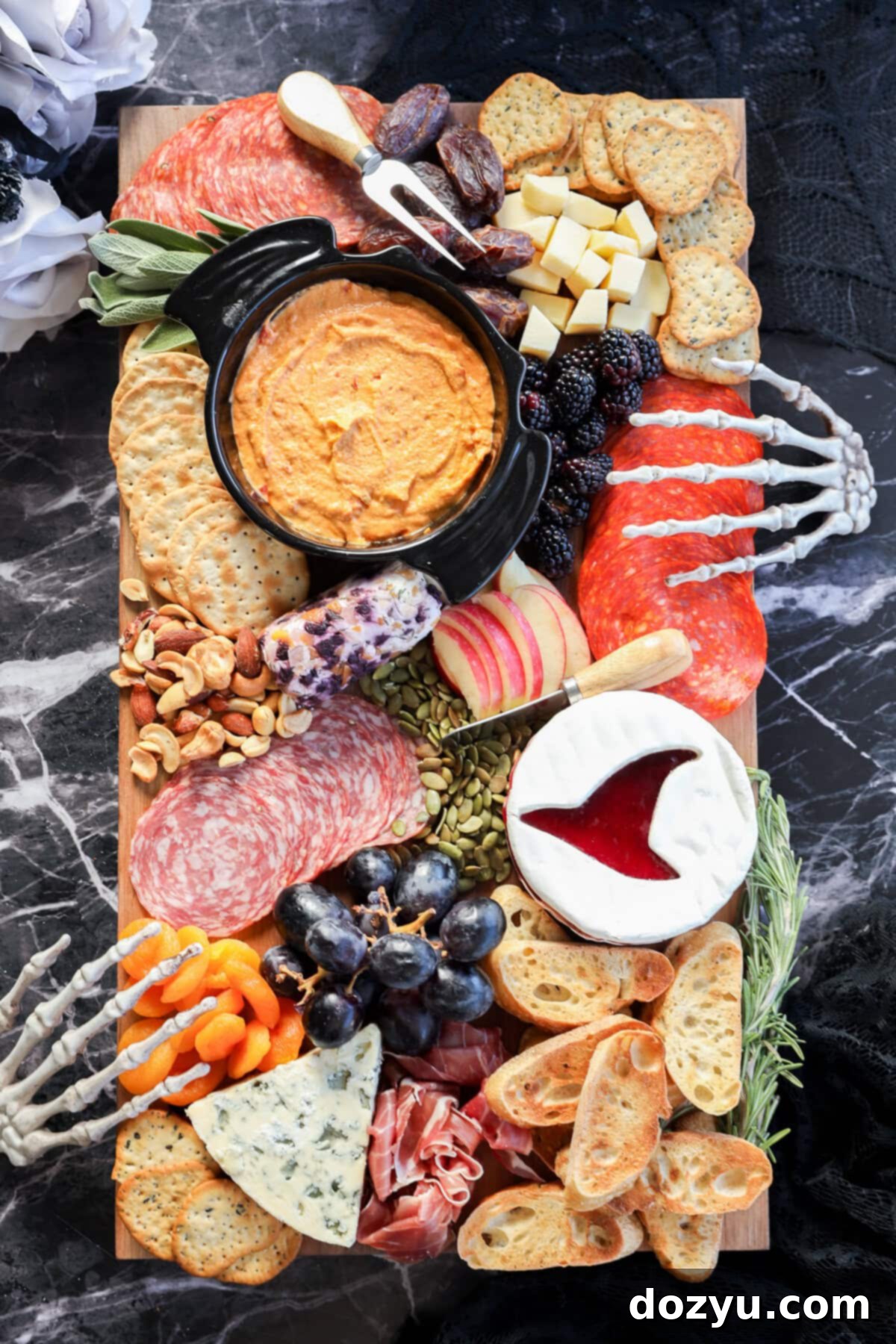 A Halloween charcuterie board featuring meats, cheeses, crackers, fruits, dips, nuts, and bones is artfully arranged with decorative skeleton hands on a dark marble surface for a spooky touch.