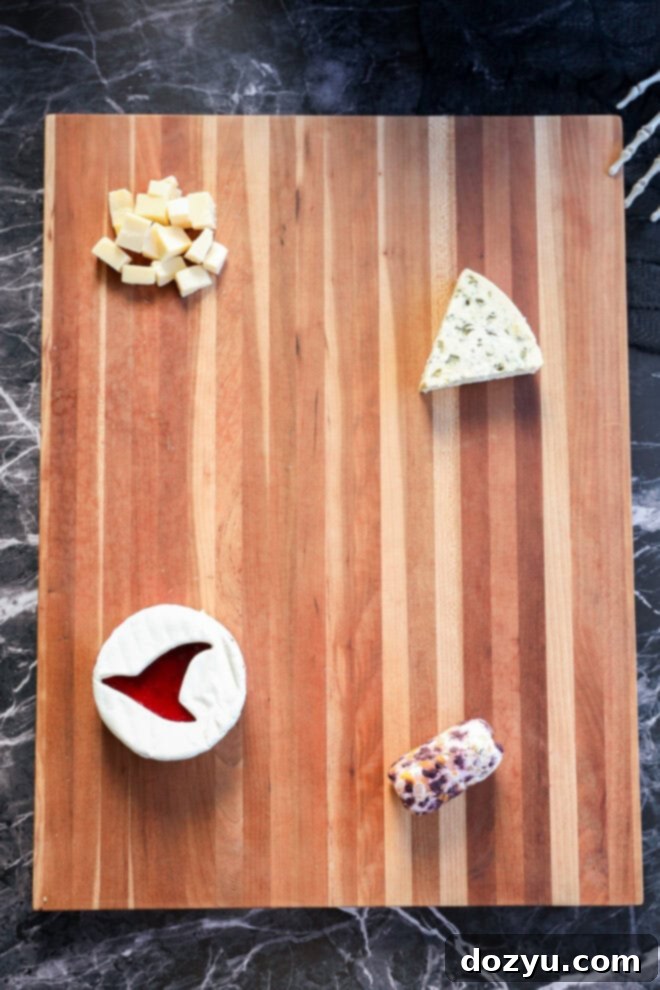 A wooden cutting board displays a halloween charcuterie board with four cheeses: cubed cheese in the top left, blue cheese wedge on the right, round brie with a slice cut out at the bottom left, and an herbed cheese log at the bottom right.
