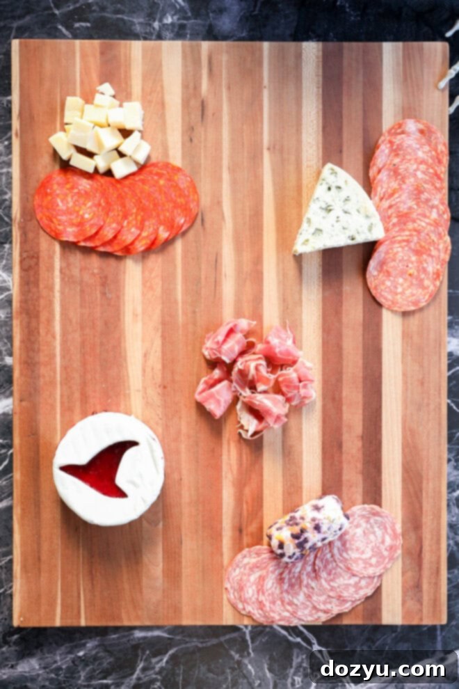 A wooden Halloween charcuterie board features sliced cured meats, cubed cheese, a wedge of blue cheese, a round of brie with jam, a log of goat cheese, and prosciutto artfully arranged on a dark marble surface.