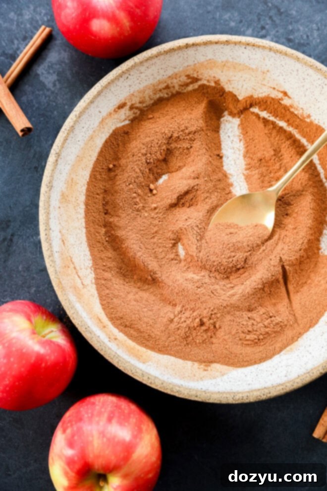 A beautifully arranged bowl of homemade apple pie spice, with a golden spoon resting in it, surrounded by vibrant red apples and rustic cinnamon sticks on a rich, dark wooden surface. The scene evokes warmth and autumn flavors, highlighting the quality of the spice blend.