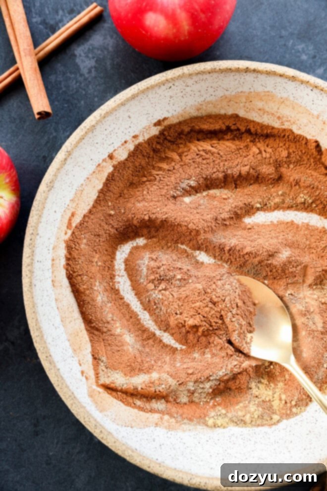 A golden spoon stirs a rich, brown mixture of ground cinnamon and other warm spices in a ceramic bowl, preparing a batch of homemade apple pie spice. The dark countertop setting is adorned with fresh red apples and fragrant cinnamon sticks, emphasizing the natural ingredients.