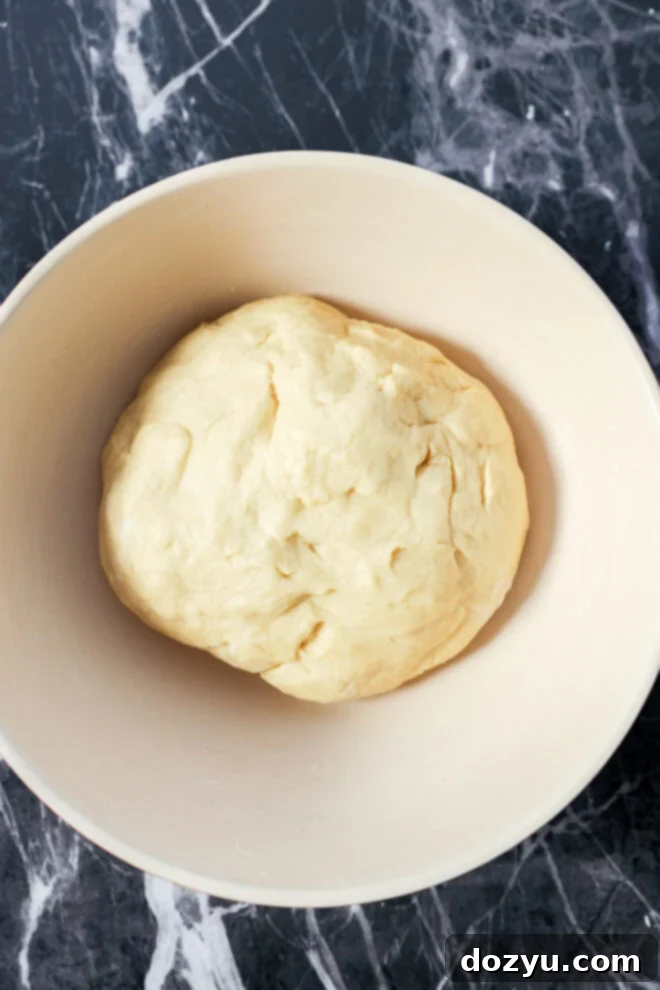 A ball of dough for Apple Pie Filled Cinnamon Rolls rests in a light-colored mixing bowl, placed on a dark, marbled countertop.