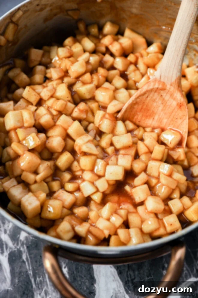 A saucepan filled with diced apples coated in a glossy cinnamon sauce, reminiscent of Apple Pie Filled Cinnamon Rolls, is being stirred with a wooden spoon on a marble surface.