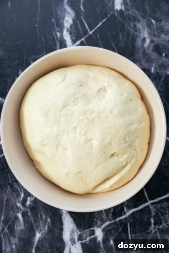 A large, smooth ball of dough—destined for Apple Pie Filled Cinnamon Rolls—rests in a light-colored bowl on a dark, marbled countertop.