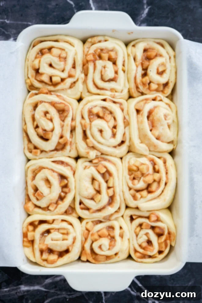 A white baking dish lined with parchment paper holds twelve unbaked Apple Pie Filled Cinnamon Rolls filled with diced apples and cinnamon, arranged in neat rows on a dark marble surface.