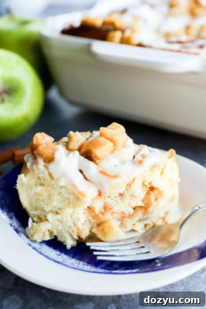 A close-up of Apple Pie Filled Cinnamon Rolls topped with diced apples and white icing on a blue and white plate with a fork. A baking dish and green apples are visible in the background.