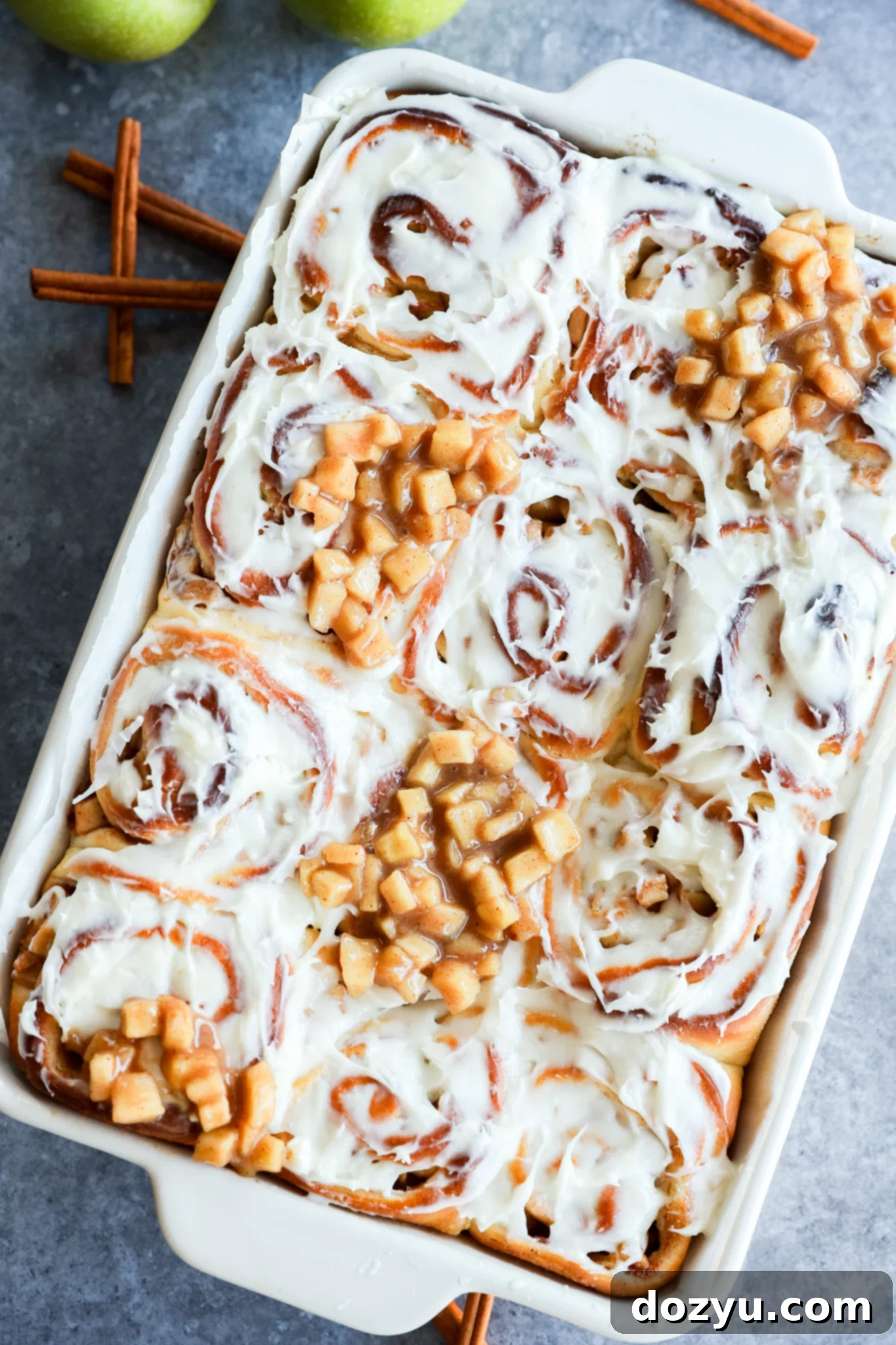 A baking dish filled with Apple Pie Filled Cinnamon Rolls, some topped with small diced apple pieces. Two green apples and cinnamon sticks are in the background on a gray surface.