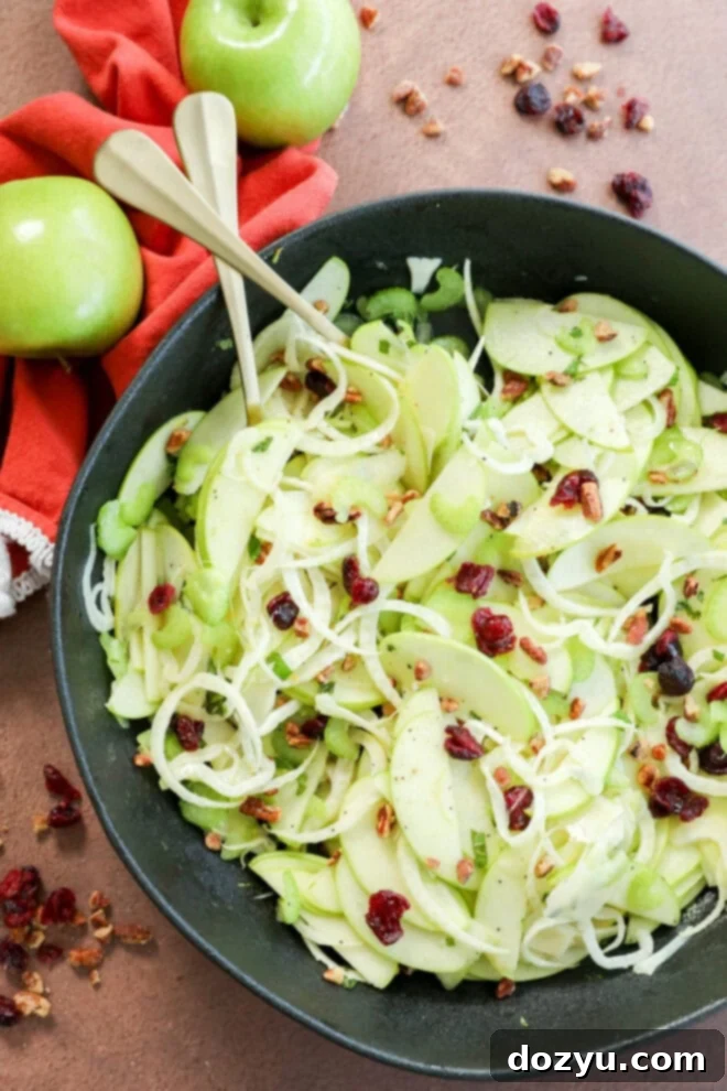 A large, inviting bowl of fresh Apple Fennel Salad, garnished with thinly sliced green apples, finely shredded cabbage, crisp celery, sweet dried cranberries, and an assortment of chopped nuts. Two elegant serving utensils are nestled in the salad, with whole green apples and a red linen napkin creating a warm, festive backdrop, perfect for a holiday meal.
