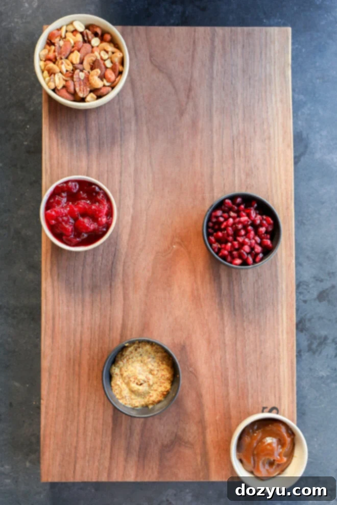 Five small bowls filled with mixed nuts, cranberry sauce, pomegranate seeds, whole grain mustard, and caramel sauce arranged on a wooden board for a Thanksgiving charcuterie board.
