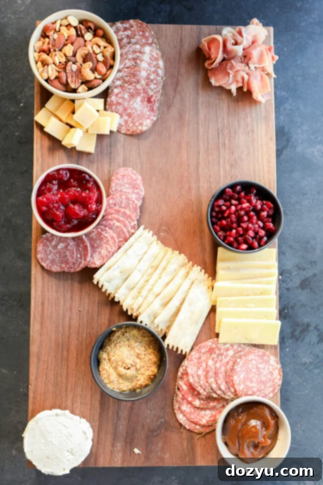 A Thanksgiving charcuterie board featuring assorted crackers, salami, cheeses, nuts, cranberry sauce, pomegranate seeds, prosciutto, mustard, goat cheese, and a small bowl of brown chutney.