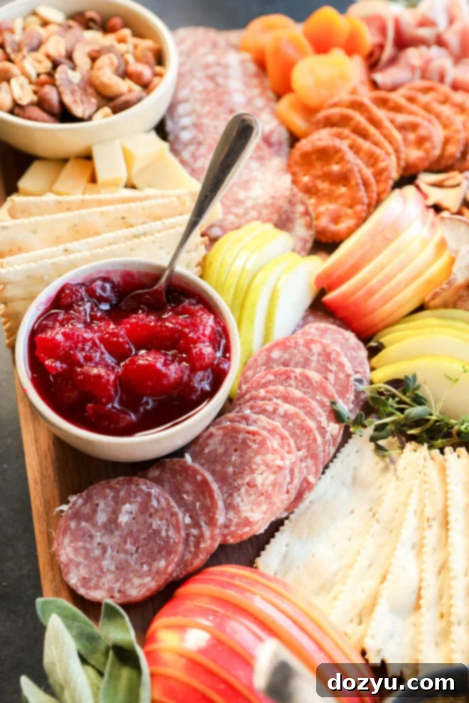 A Thanksgiving charcuterie board with salami, apples, pears, crackers, dried apricots, mixed nuts, and a bowl of red fruit jam.