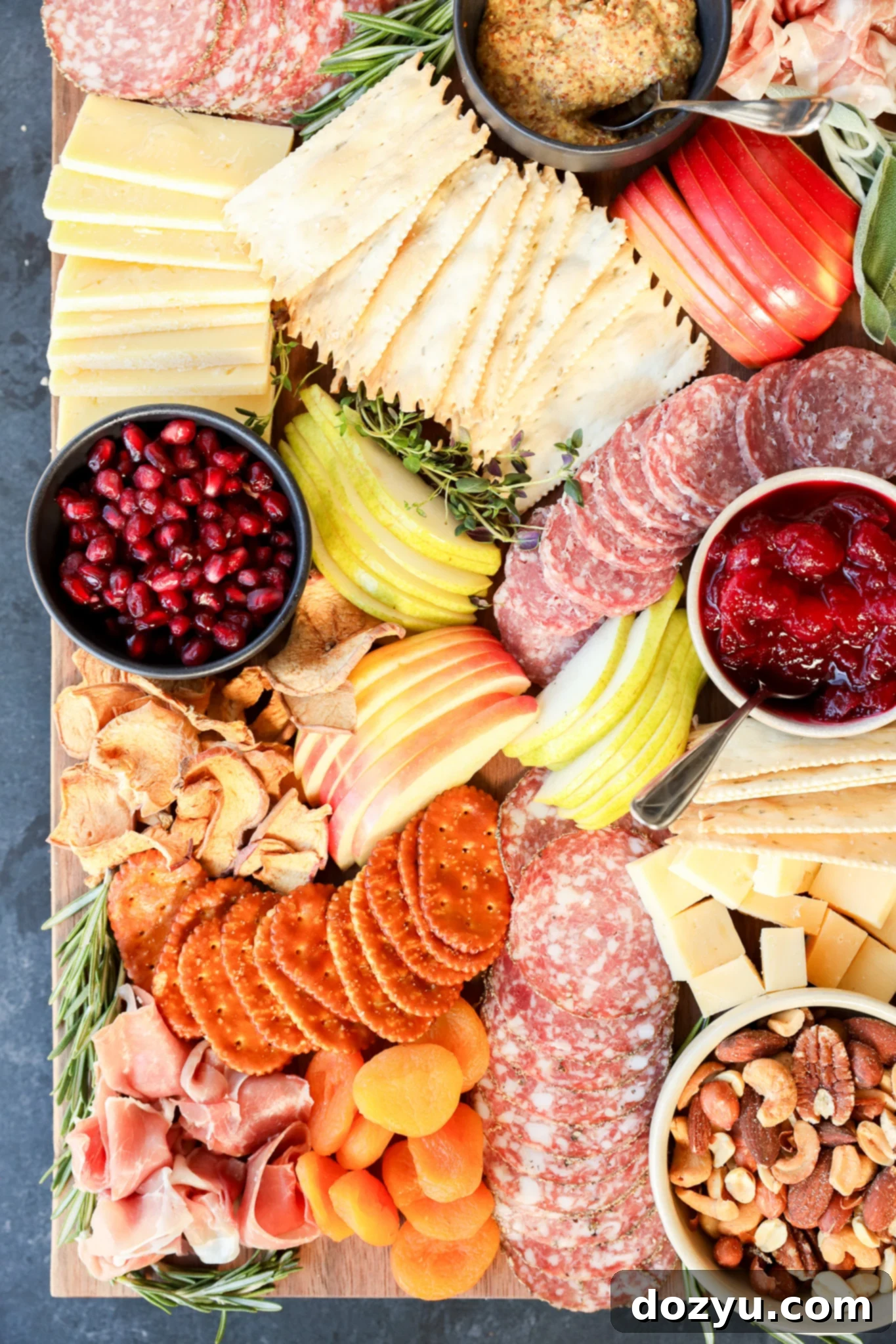 A Thanksgiving charcuterie board with salami, prosciutto, crackers, cheeses, apple and pear slices, dried apricots, nuts, pomegranate seeds, dried fruit chips, mustard, cranberry sauce, and herb garnishes.