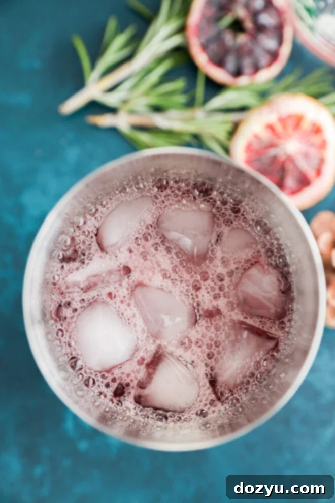 A close-up top view of a glass filled with a fizzy purple christmas vodka cocktail and ice cubes, set on a blue surface, with blood orange slices and sprigs of rosemary in the background.