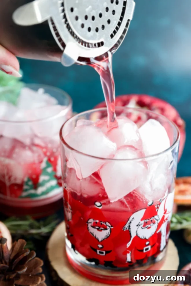 A close-up of a festive glass decorated with Santa Claus illustrations, filled with ice, as a pink-red christmas vodka cocktail is poured in using a shaker. Another similar glass and holiday décor appear in the background.