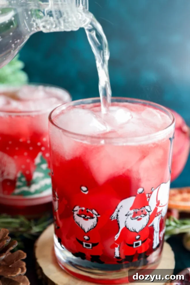 A clear liquid is being poured into a glass filled with ice and a bright red christmas vodka cocktail. The glass is decorated with images of Santa Claus, with another festive glass and decorations in the background.