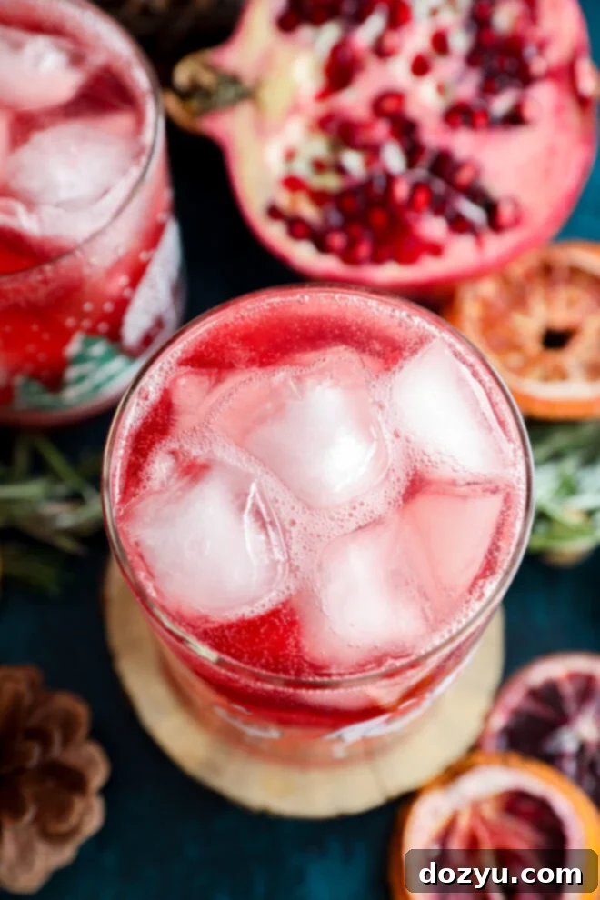A close-up of a glass filled with a red, ice-filled christmas vodka cocktail, surrounded by pomegranate halves, dried orange slices, pinecones, and sprigs of rosemary on a dark background.