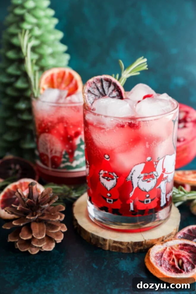 A festive christmas vodka cocktail with ice, garnished with dried citrus and rosemary, served in a Santa Claus glass. Nearby sit a second similar drink, a pinecone, wooden coaster, dried fruit, and a green tree-shaped candle.