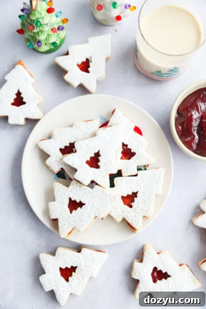 A plate of holiday cookies dusted with powdered sugar, filled with red jam, sits on a table. Nearby are more cookies, a glass of milk, a bowl of jam, and a small ceramic Christmas tree decoration.