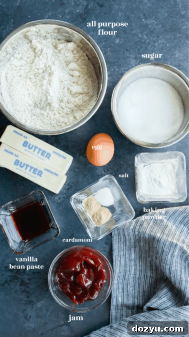Top-down view of baking ingredients for christmas tree cookies on a dark surface: flour, sugar, two butter sticks, an egg, salt, baking powder, cardamom, vanilla bean paste, and a bowl of jam, with a striped kitchen towel beside them.