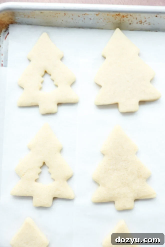 Christmas tree-shaped sugar cookies, some with small tree cutouts in the center, are arranged on a baking sheet lined with parchment paper.