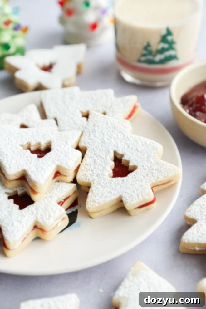 A plate of Christmas tree-shaped linzer cookies dusted with powdered sugar, filled with red jam. A glass with a festive tree design and a bowl of jam are in the background.
