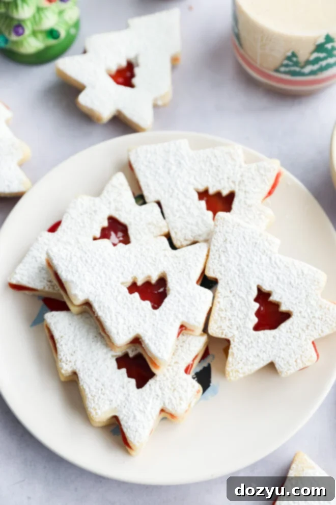A plate of baked goodies dusted with powdered sugar, each with a cutout tree in the center revealing red jam filling, surrounded by festive decor.