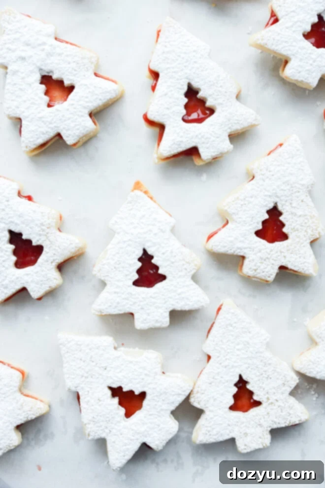 Christmas tree cookies dusted with powdered sugar, each with a small tree-shaped cutout in the center showing red jam filling underneath. The cookies are arranged on a light surface.
