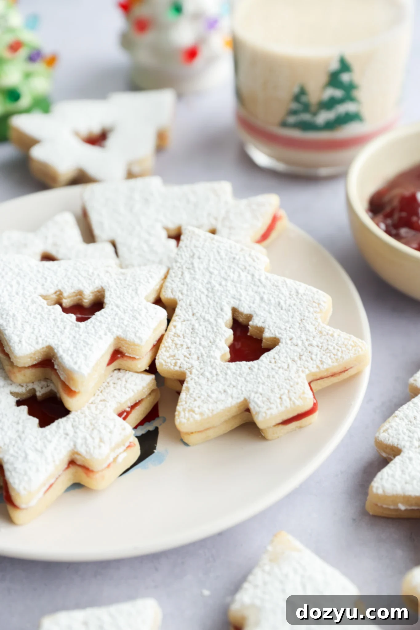 A plate of Christmas tree-shaped linzer cookies dusted with powdered sugar, filled with red jam. A glass with a festive tree design and a bowl of jam are in the background.