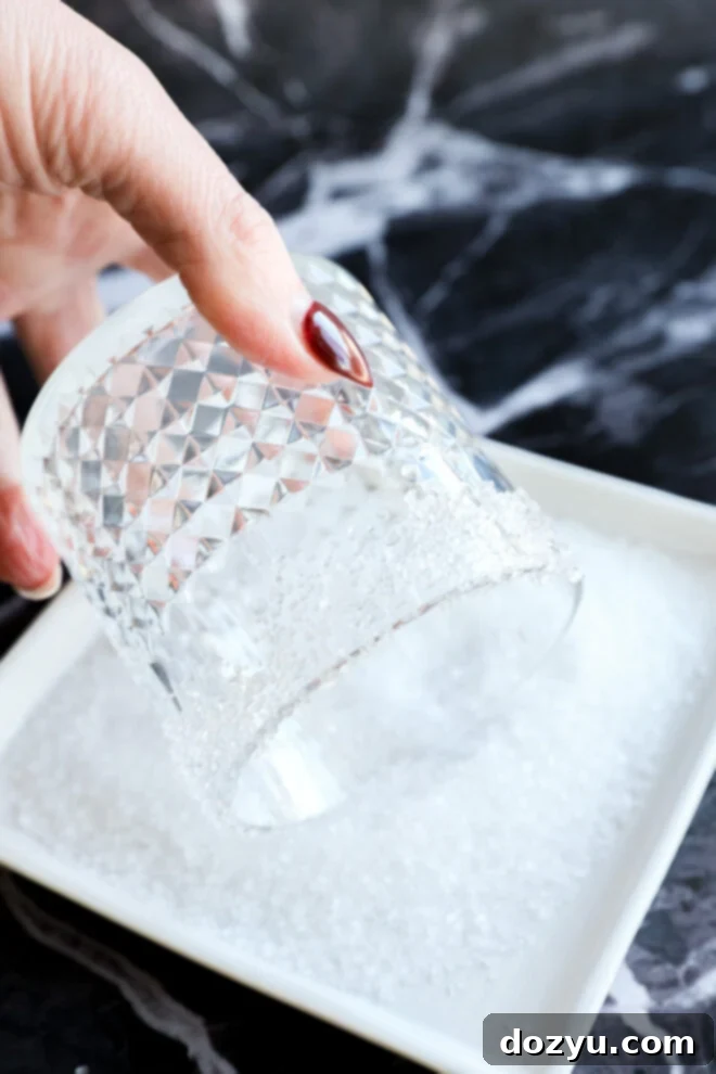 A hand dips the rim of a textured glass into coarse white salt. The background is a dark, marbled surface.