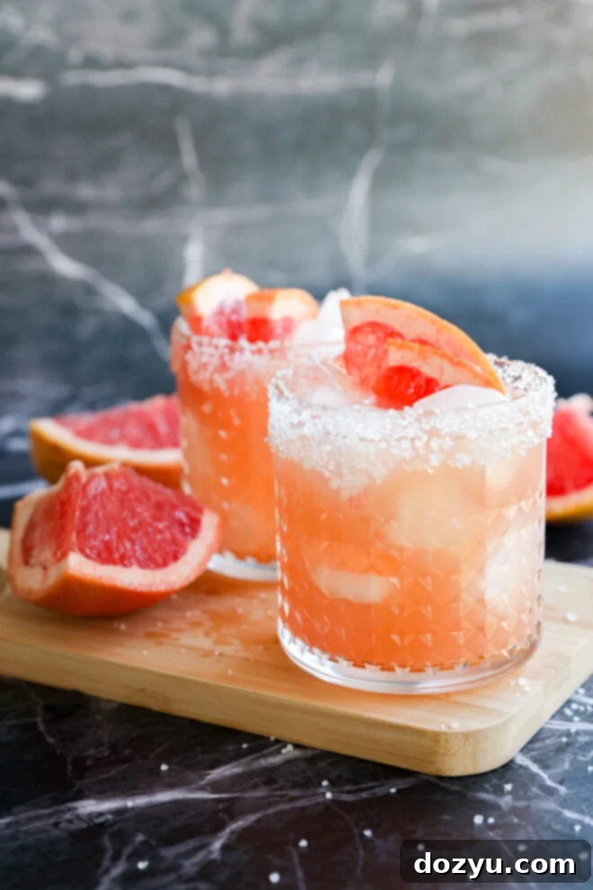 Two glasses of Grapefruit Vodka Cocktail with ice and salted rims sit on a wooden board, garnished with grapefruit wedges. Fresh grapefruit halves are nearby, all set against a marble background.