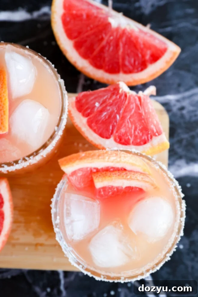 Two glasses of Grapefruit Vodka Cocktail with ice and salted rims, garnished with grapefruit slices. Fresh grapefruit wedges are placed nearby on a dark marble surface and a wooden board.